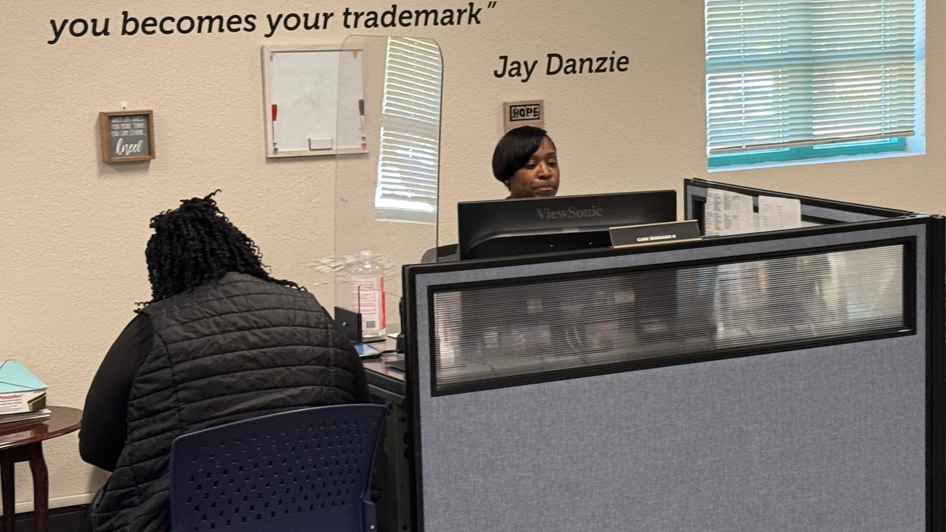 Woman in face mask behind front desk of Housing Navigation Center & back of woman in front of desk