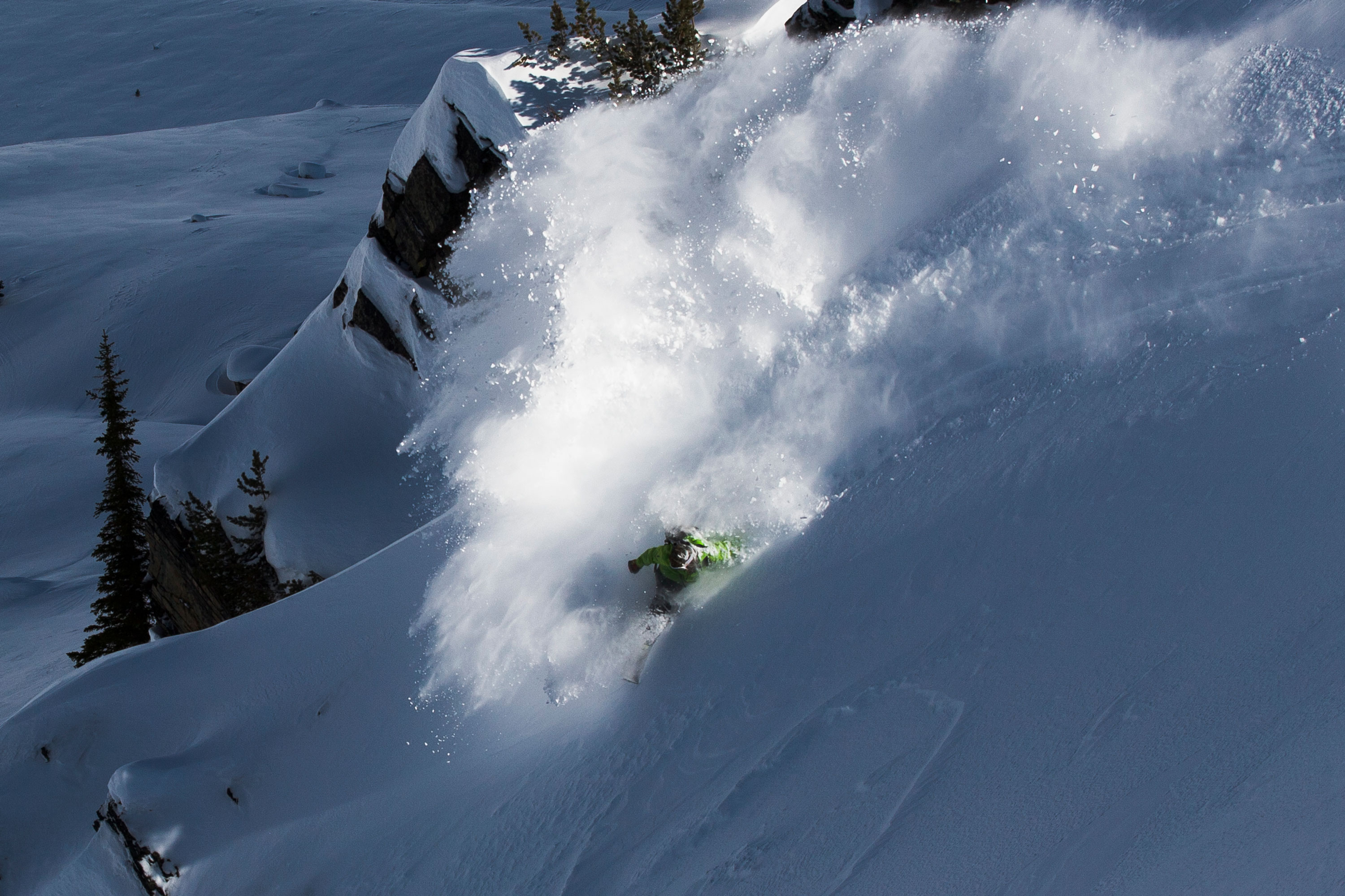 Skier in a green jacket skiing downhill through deep powder snow, creating a large cloud of snow spray.