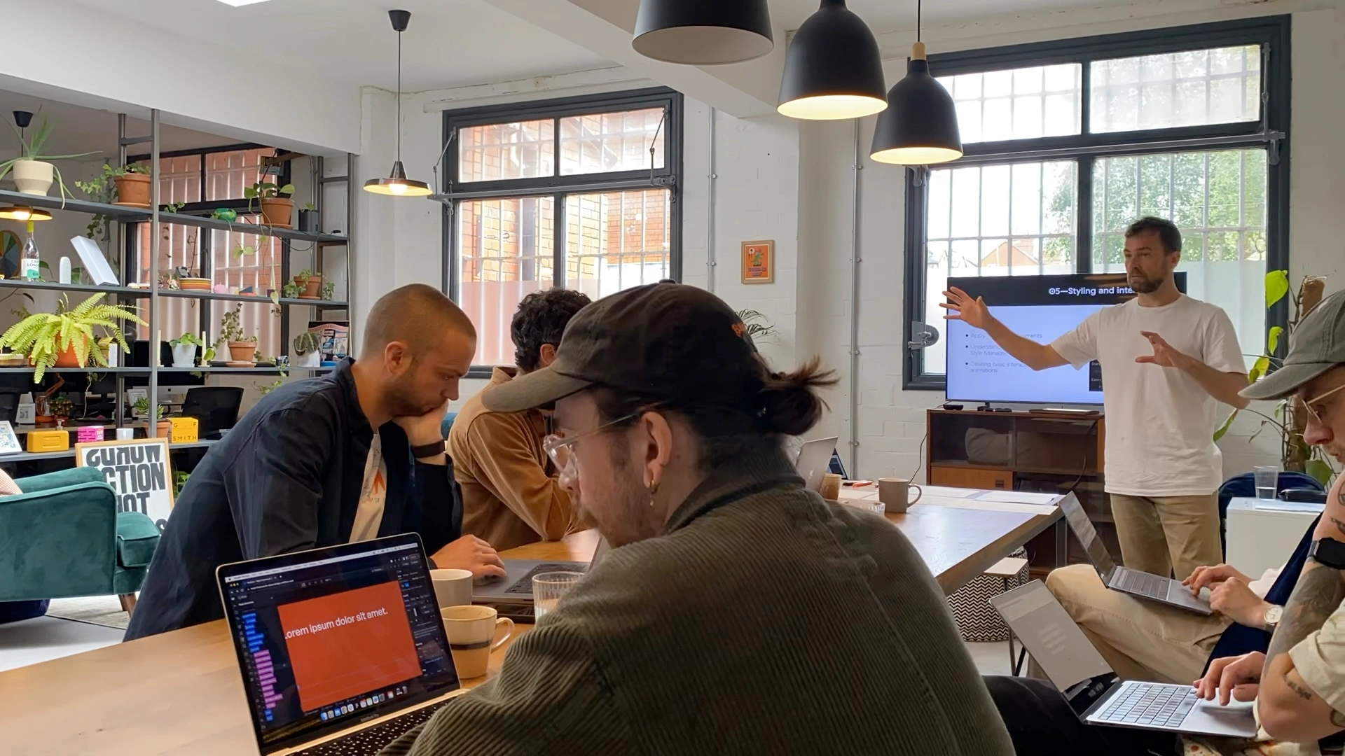 Group of people in a modern office attending a workshop with a man presenting near a screen and others working on laptops at a large table.