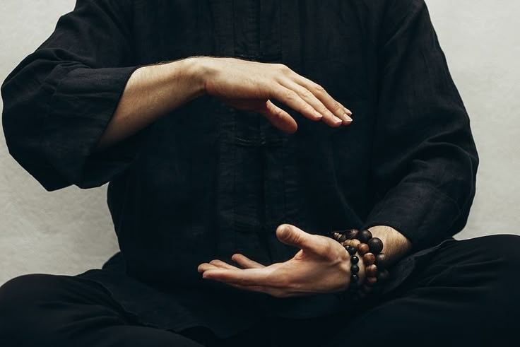 Person in black clothing sitting cross-legged, holding hands in a meditative pose with wooden bead bracelets on the wrist.