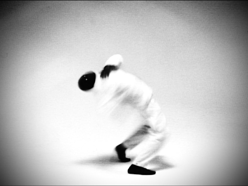 Blurred black and white photo of a person in motion wearing loose white clothing and a dark cap, bending backwards.