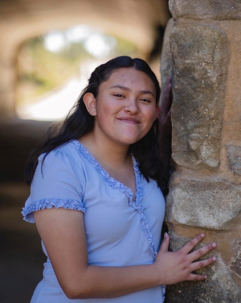 A picture of Ahtziri. She is holding a brick wall to the right. She is smiling and is wearing a light blue dress.