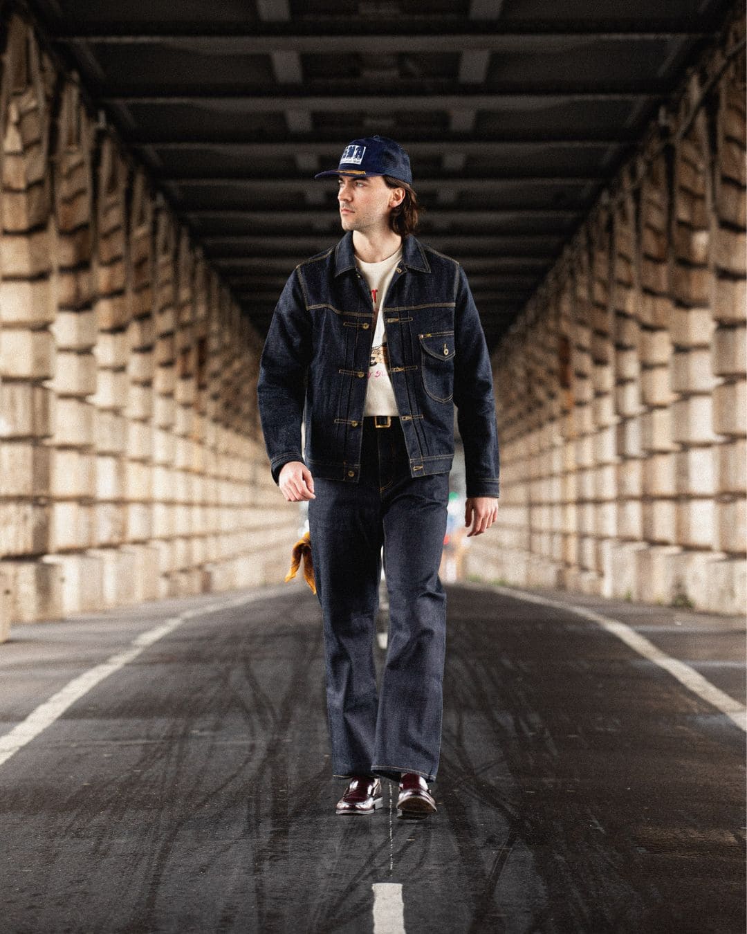 Modern Americana full denim look with trucker jacket, navy cap and burgundy loafers, walking under an urban bridge