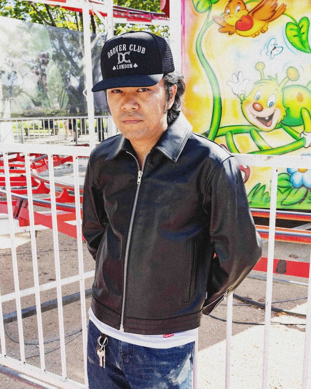 Modern Americana look with black leather jacket and Drover Club trucker cap in front of a colorful funfair backdrop