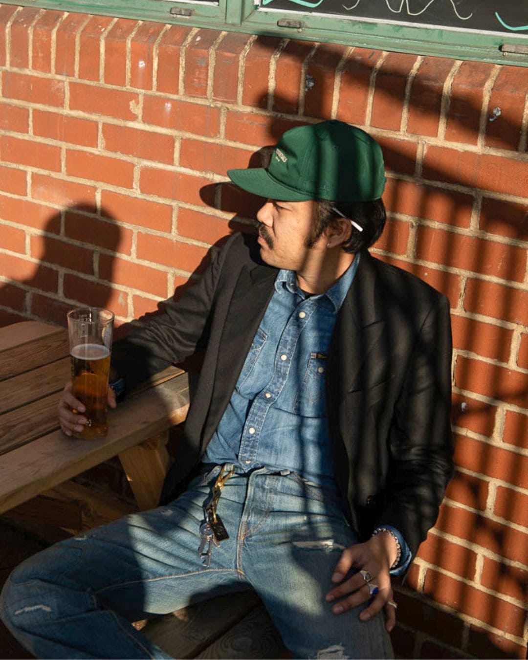 Modern Americana look with black blazer, light denim and green cap, relaxing on a sunny terrace with a beer