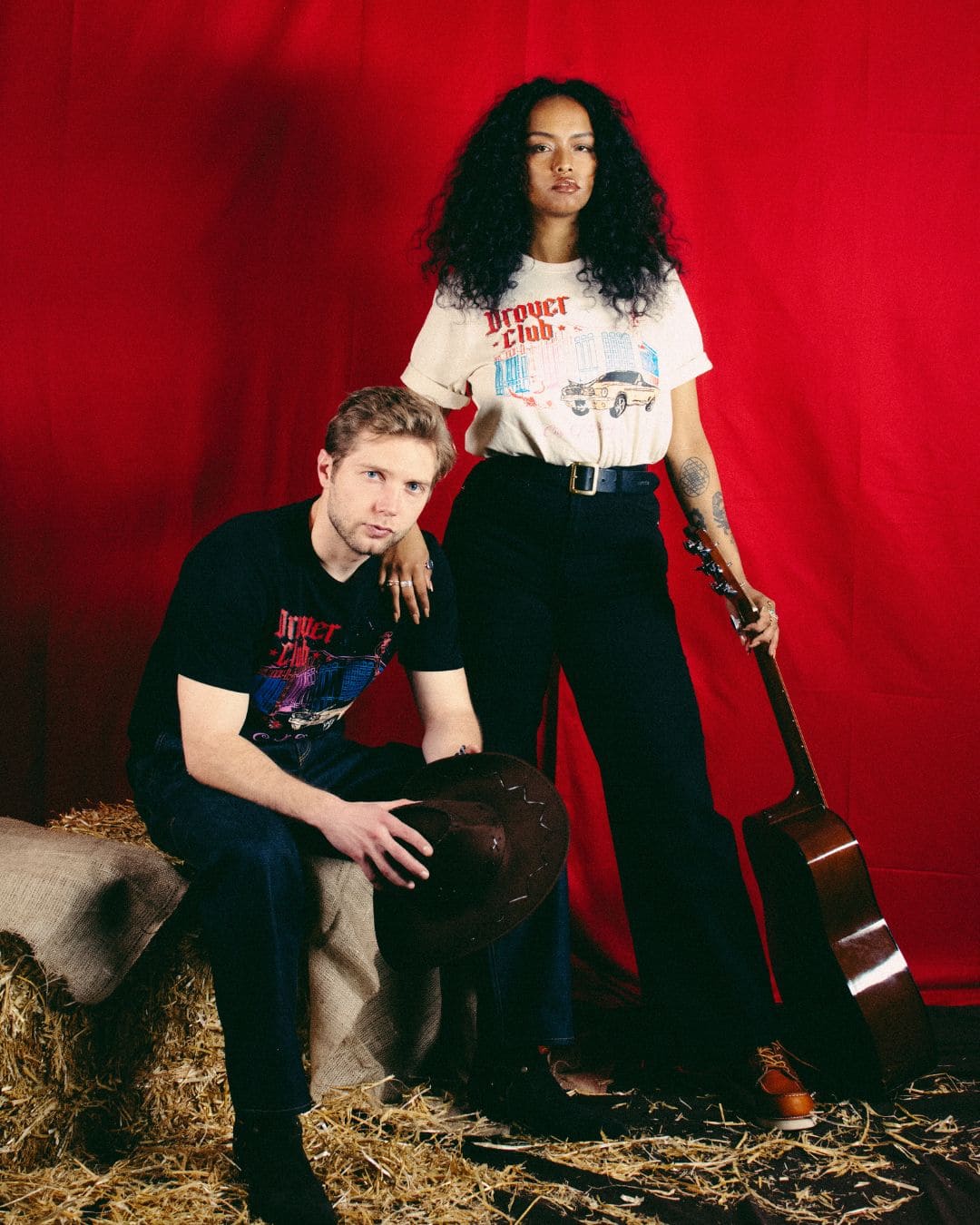 Modern Americana couple look with matching Drover Club graphic tees, western styling, acoustic guitar and red stage backdrop