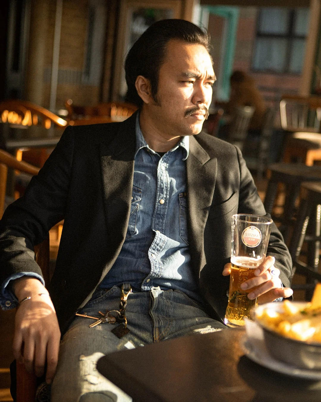 Wai Phyo, co-founder of Drover Club, seated in a bar wearing a dark blazer and denim shirt while holding a beer.