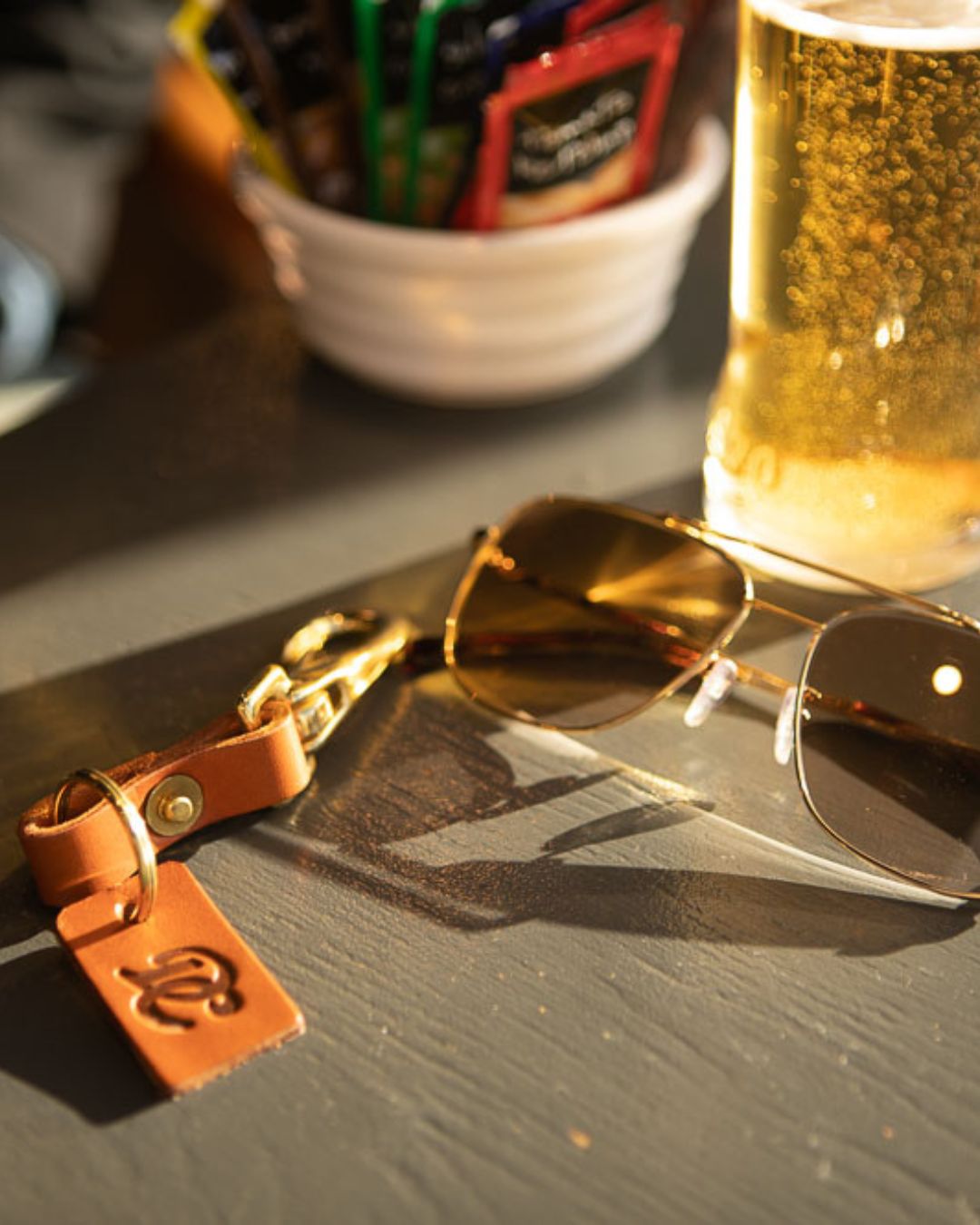 Tan Drover Club leather key clip placed on a table next to sunglasses and a glass, urban lifestyle detail.