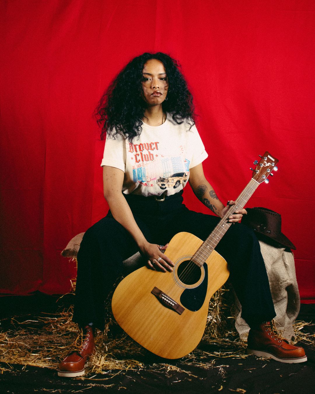 Woman in Drover Club City of Paradise tee in natural paired with black trousers and boots, sitting with guitar in Western-inspired setting