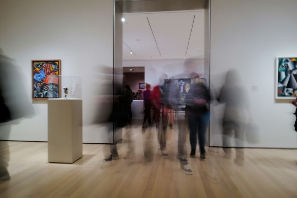 Visitors walking through a contemporary art gallery exhibition with paintings and sculptures on display.