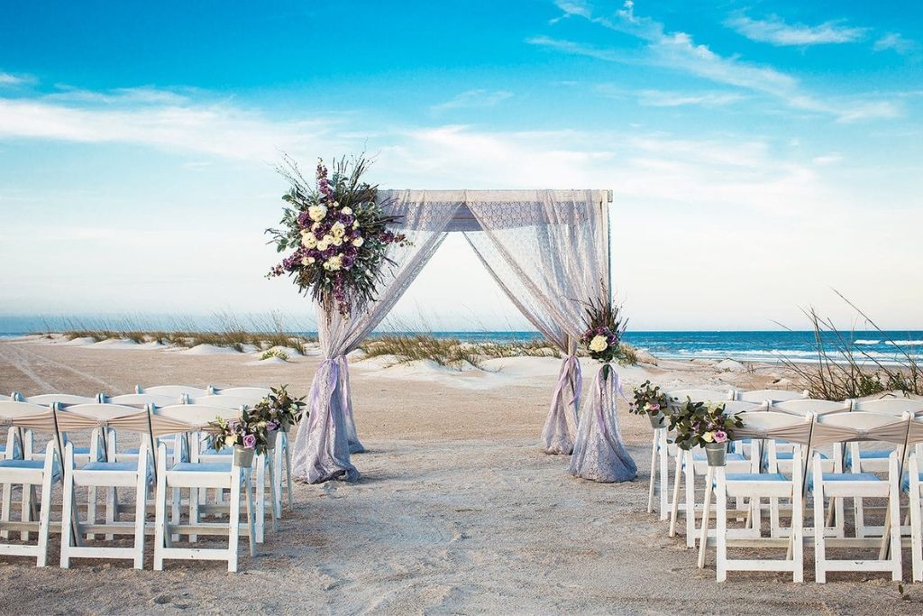 Elegant beach wedding ceremony with a floral arch overlooking the sea and rows of white chairs on the sand.