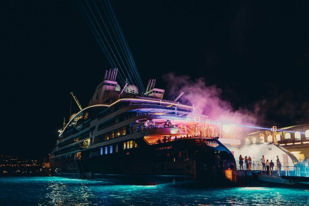 Luxury yacht hosting an evening party at a marina with guests gathering on deck under colourful lights.