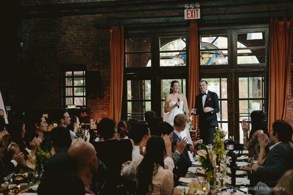 Wedding reception dinner with formally dressed guests seated at long tables while the newlyweds give a speech at an elegant indoor venue.