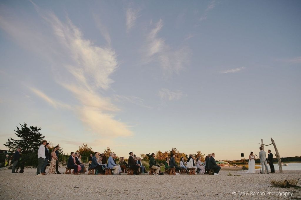 Beach wedding ceremony at sunset with formally dressed guests seated on the sand beside the water.
