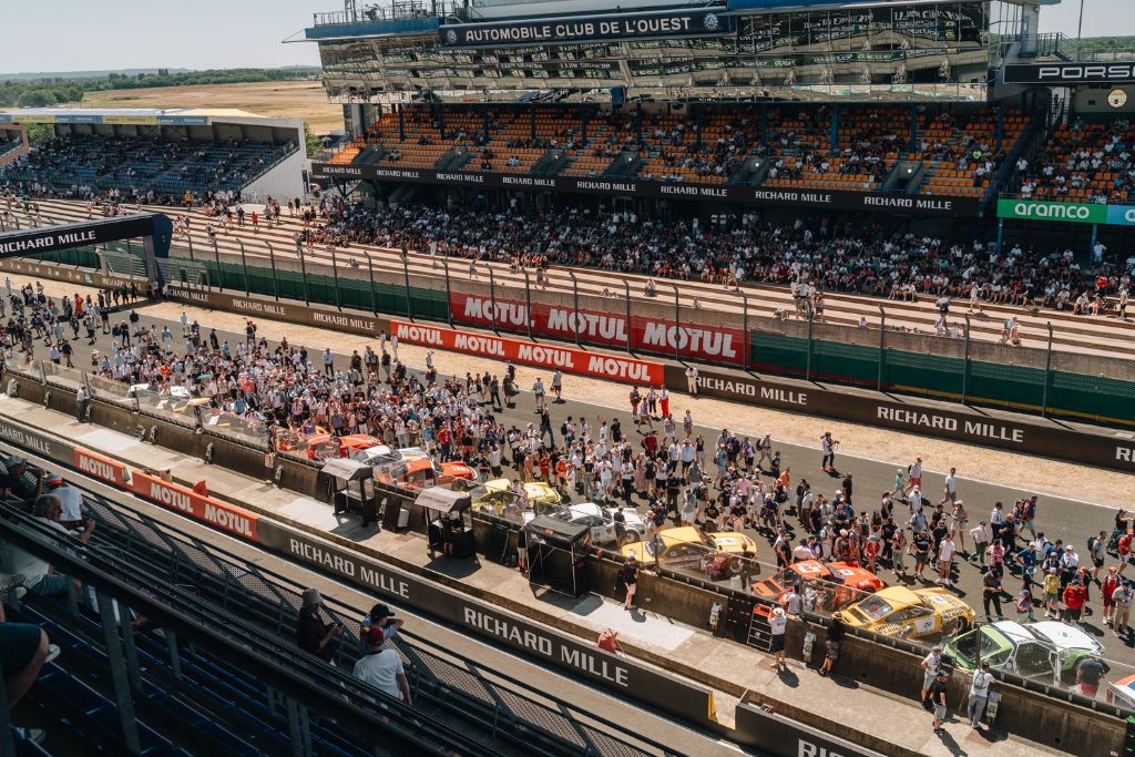 Spectators and historic race cars in the pit lane at Le Mans Classic, with the main grandstand overlooking the circuit.