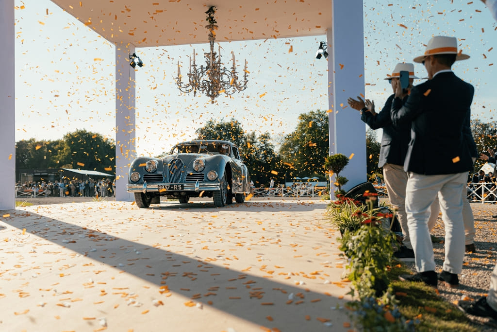 Vintage car arriving under a pavilion at Chantilly Arts & Elegance, with confetti falling and spectators watching.