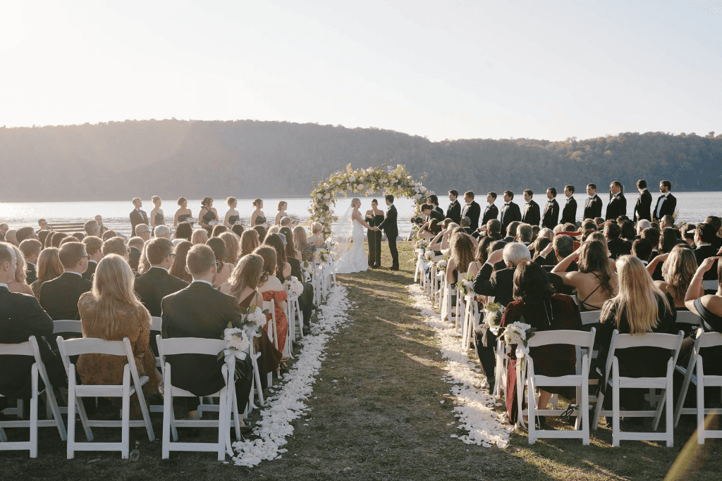 Outdoor summer wedding ceremony by a lake, with guests seated as the couple exchanges vows beneath a floral arch.