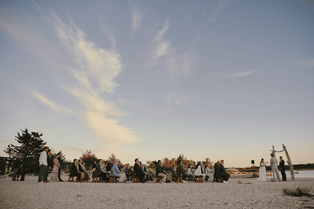 Beach wedding ceremony at sunset with formally dressed guests seated on the sand beside the water.
