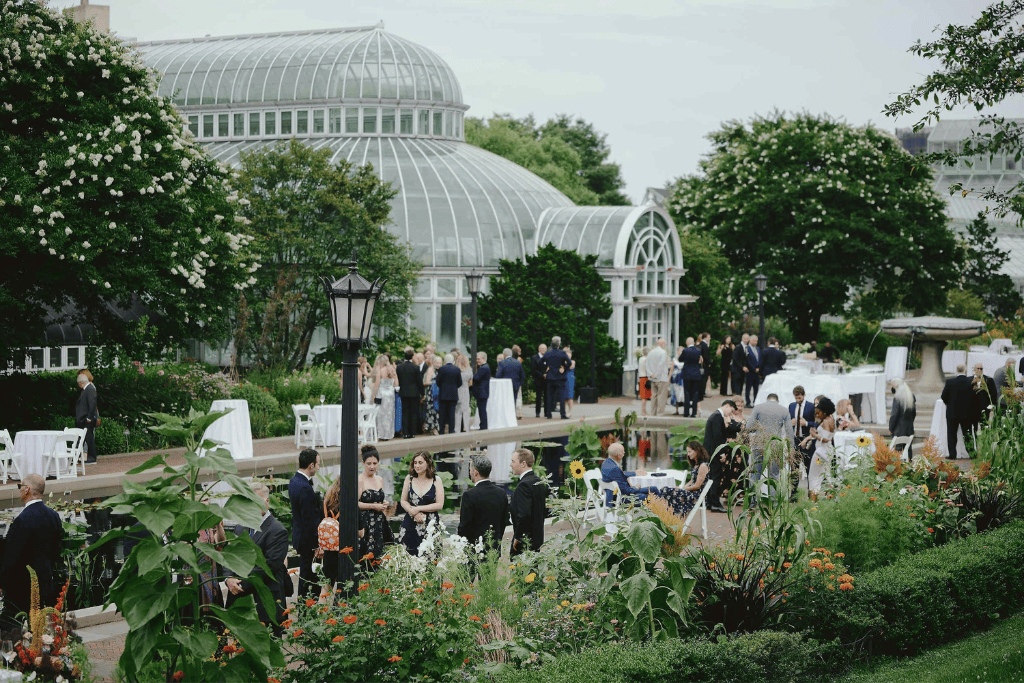 Guests gathering for an elegant garden wedding reception beside a glass conservatory surrounded by flowers and greenery.