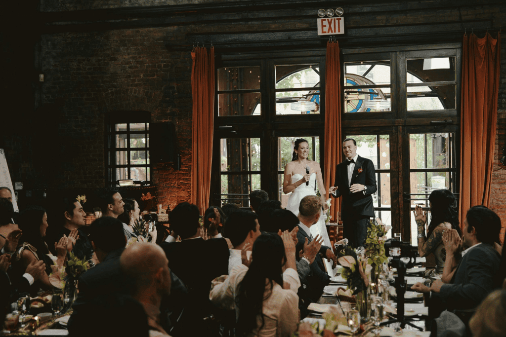 Wedding reception dinner with formally dressed guests seated at long tables while the newlyweds give a speech at an elegant indoor venue.