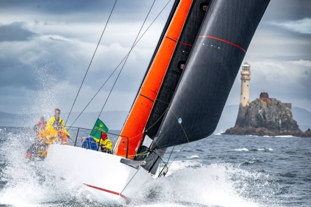 Sailing yacht racing through rough seas during the Rolex Fastnet Race, with the Fastnet Rock lighthouse in the background.