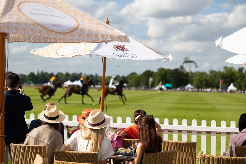 Spectators watching a polo match at Guards Polo Club from a hospitality area beside the field.