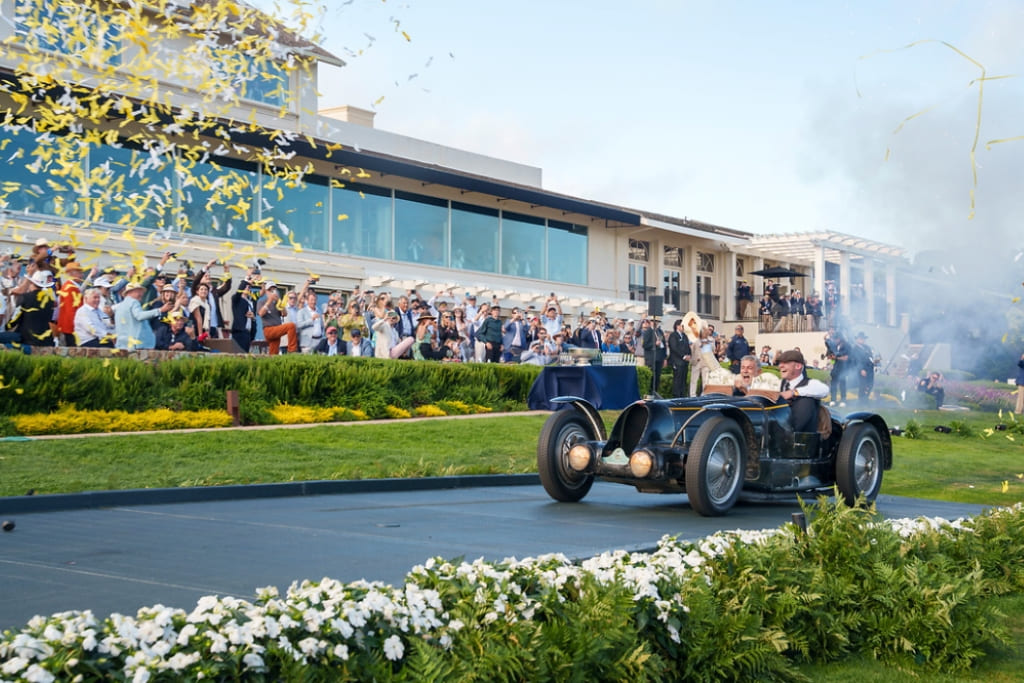 Vintage race car celebrating at the Pebble Beach Concours d’Elegance, with spectators cheering beside the lawn.