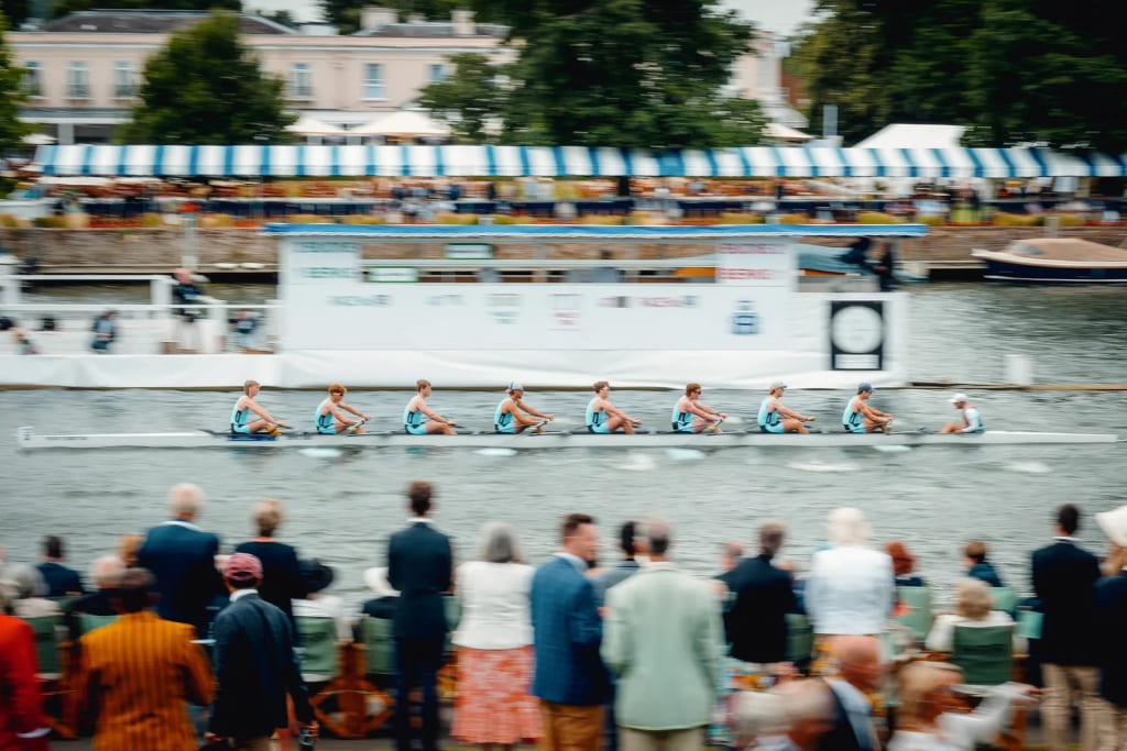 Rowing crew racing at Henley Royal Regatta, with spectators watching from the riverbank.