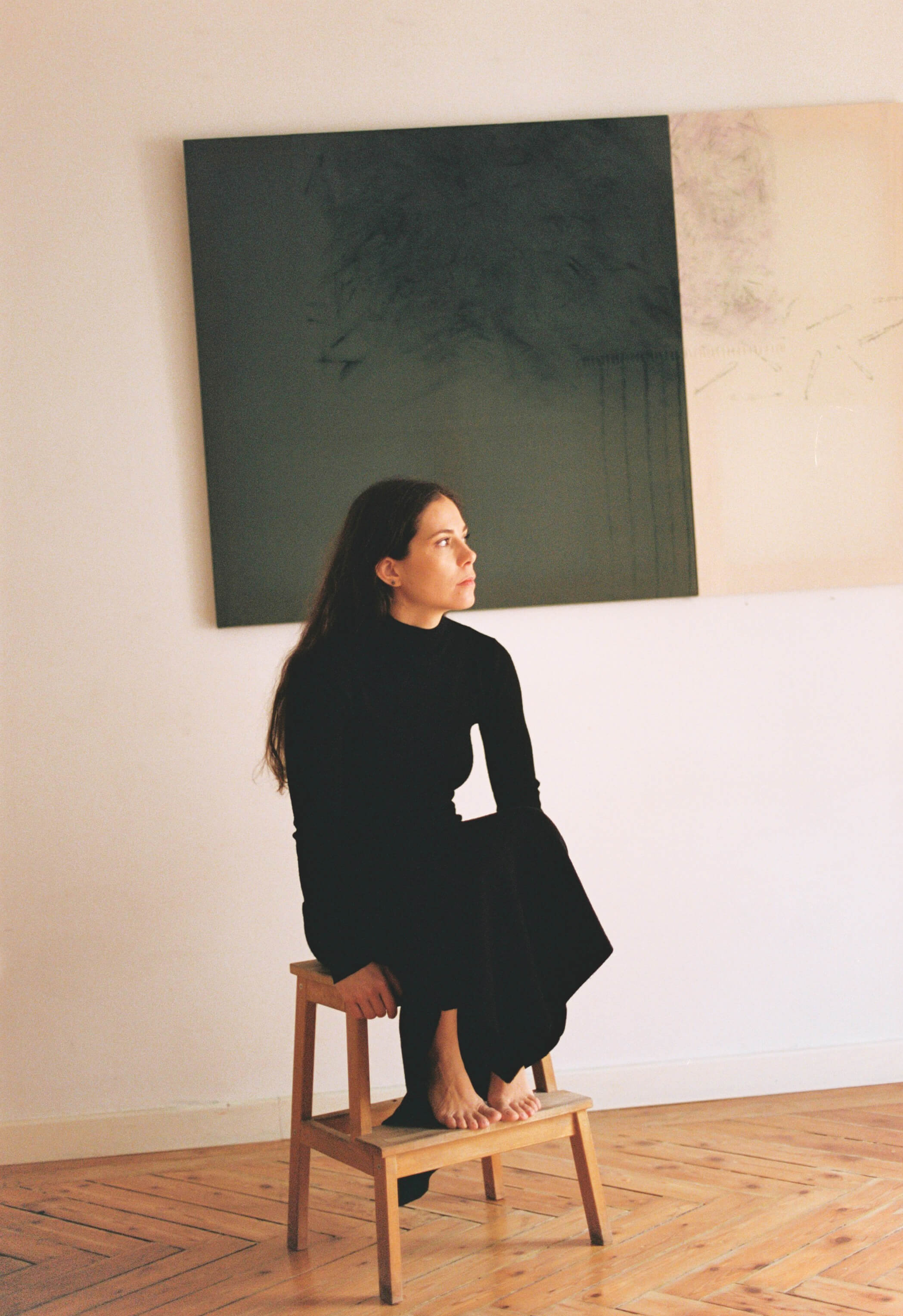 Woman in black sitting barefoot on a wooden stool in a minimalist room with wood flooring and abstract artwork on the wall.