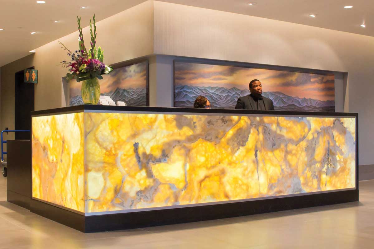 Reception desk with glowing yellow marble panels, a vase with flowers, and two people behind it in a modern lobby with mountain artwork in the background.