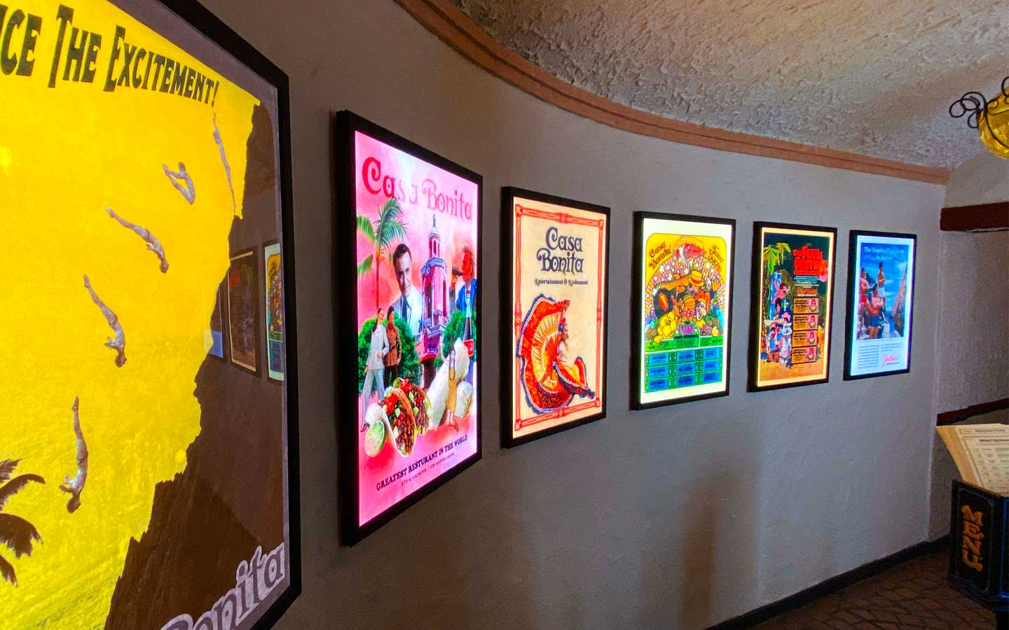 Wall displaying six colorful illuminated vintage-style posters advertising Casa Bonita restaurant with a menu stand visible on the right.