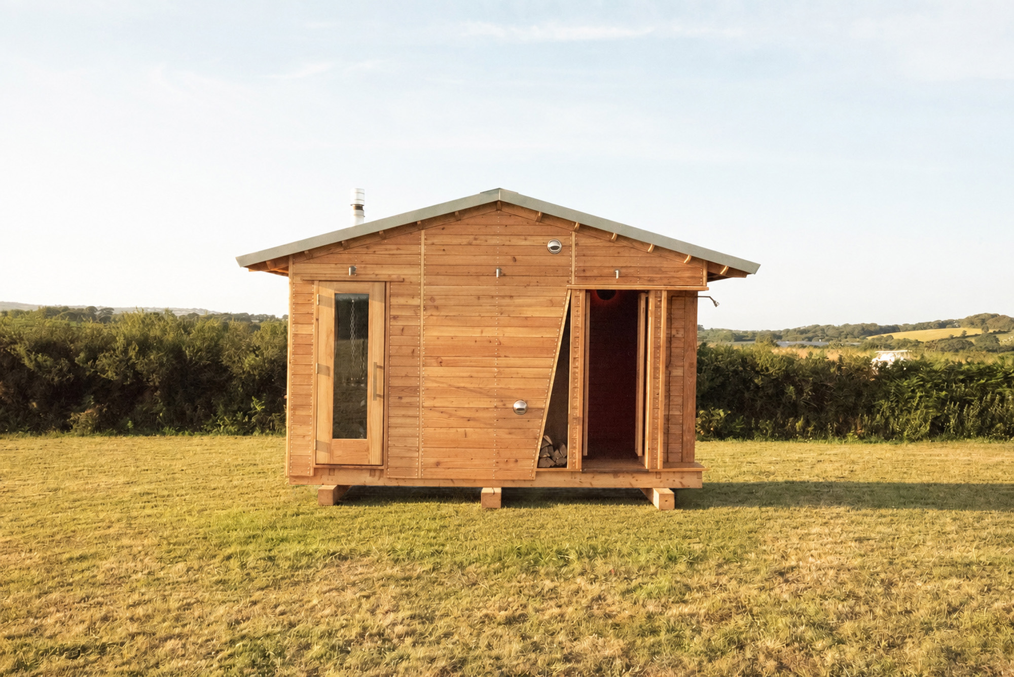 Wooden cabin with open door and window standing on grass with bushes and hills in the background.