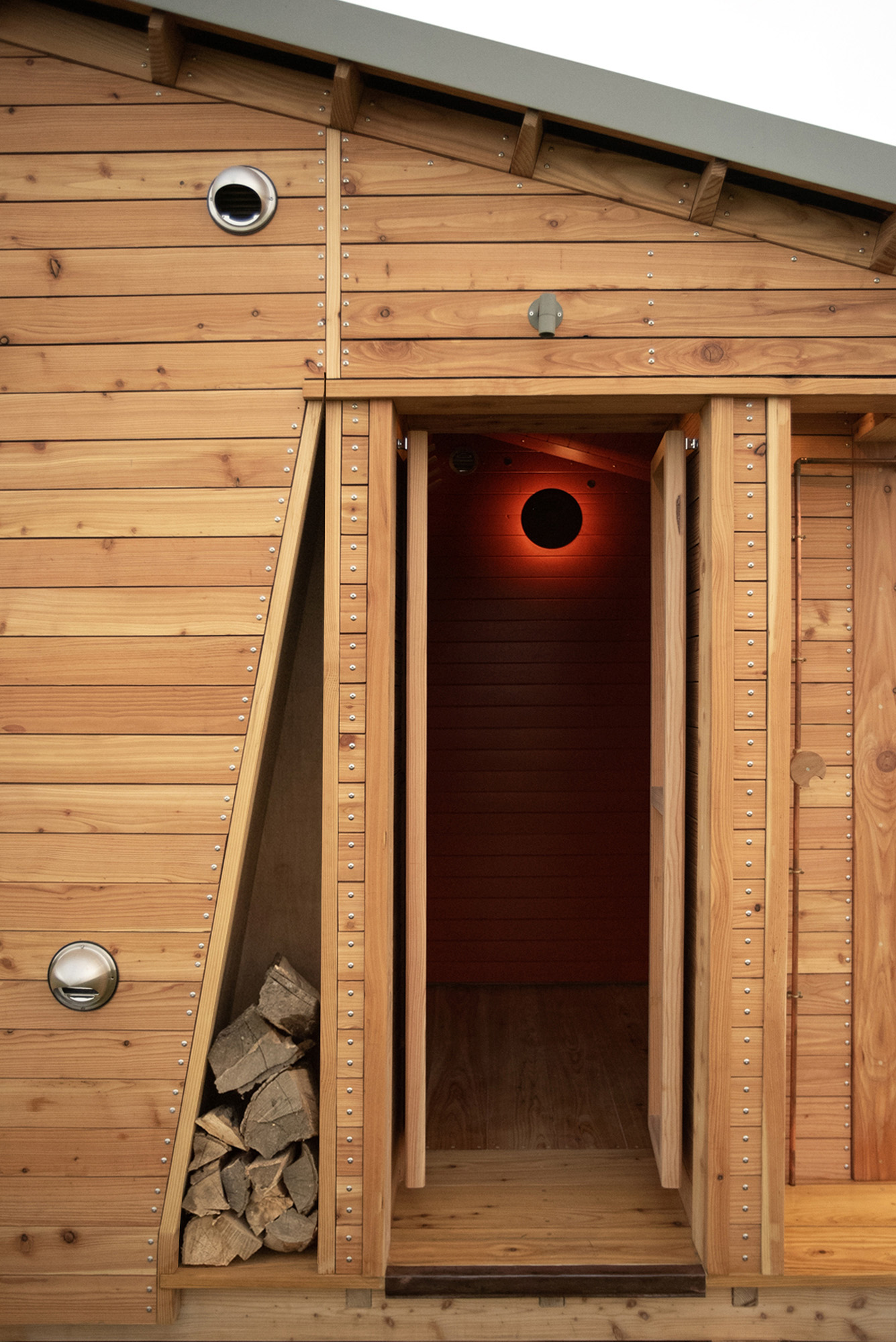 Wooden cabin door open showing interior with red light and stacked firewood on side.