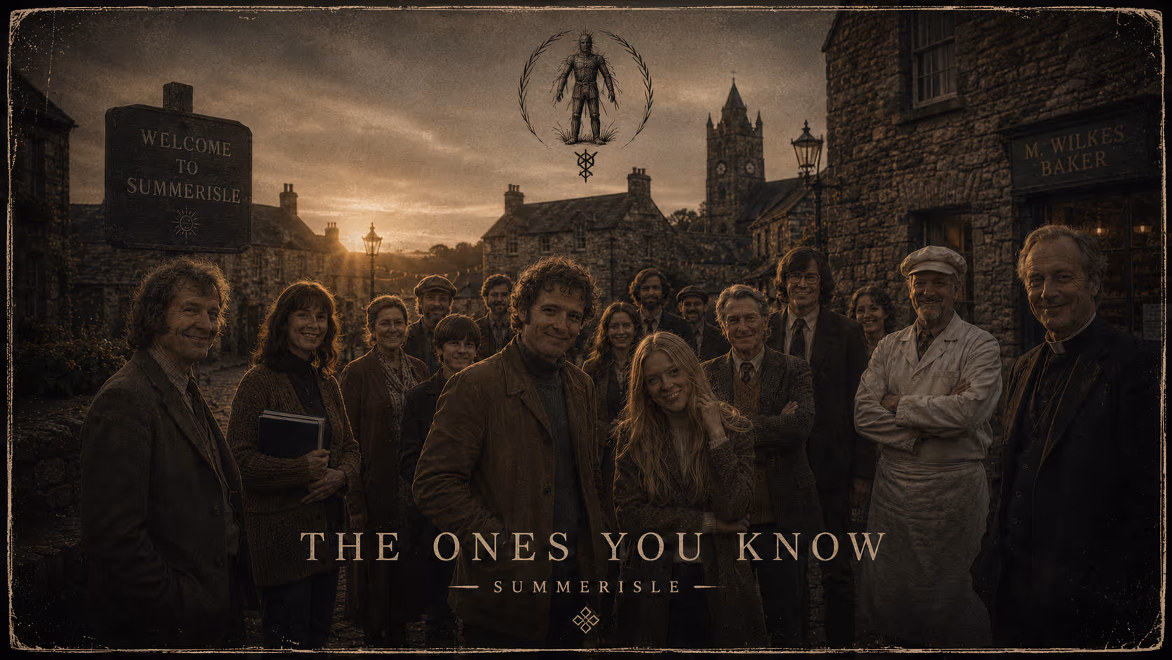 Group of people posing in an old village at sunset with signs reading Welcome to Summerisle and M. Wilkes Baker.