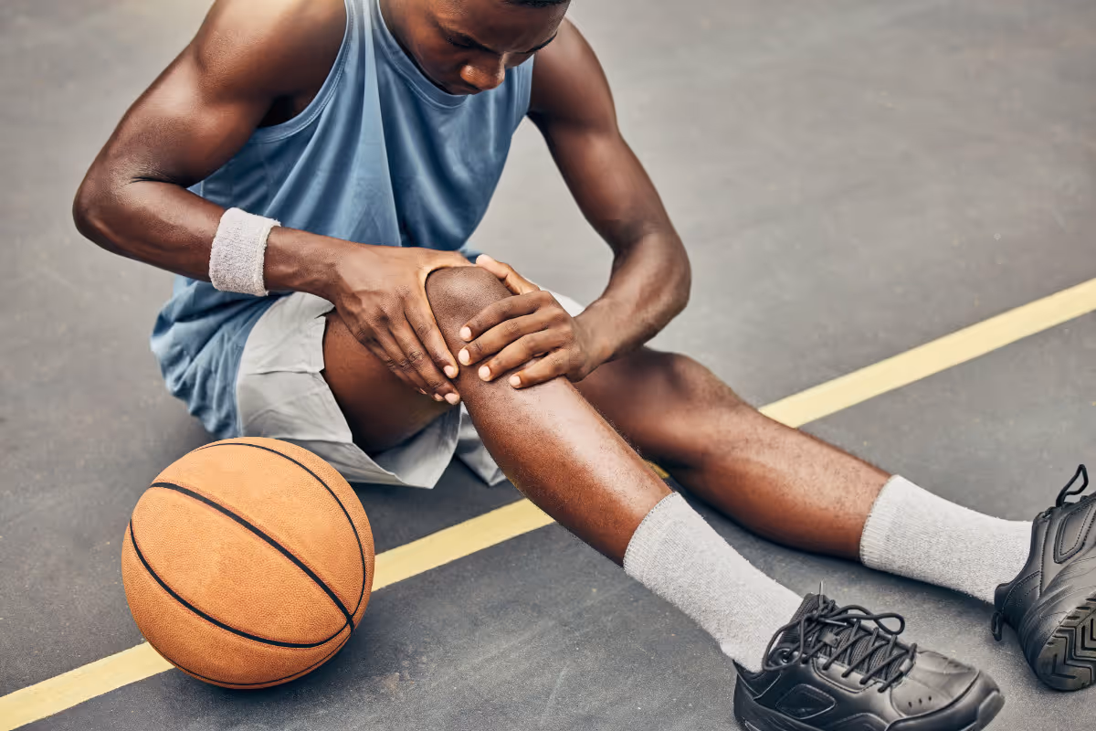 Male basketball player sitting on court holding his knee in pain next to a basketball.