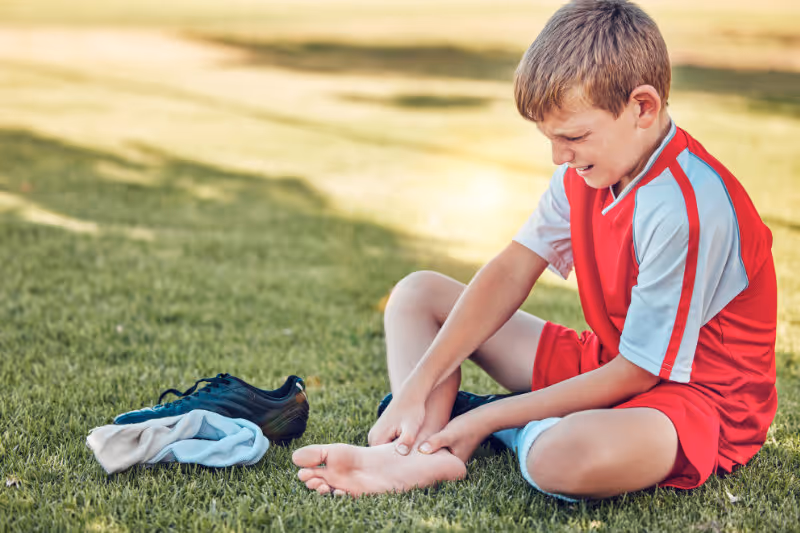 Young boy in red soccer uniform sitting on grass, holding his ankle with a pained expression, next to his soccer shoes and socks.