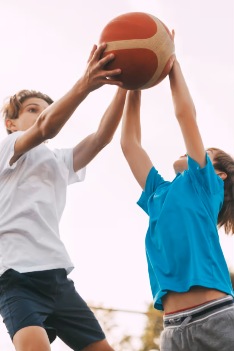 Two boys reaching up to catch a basketball outdoors.