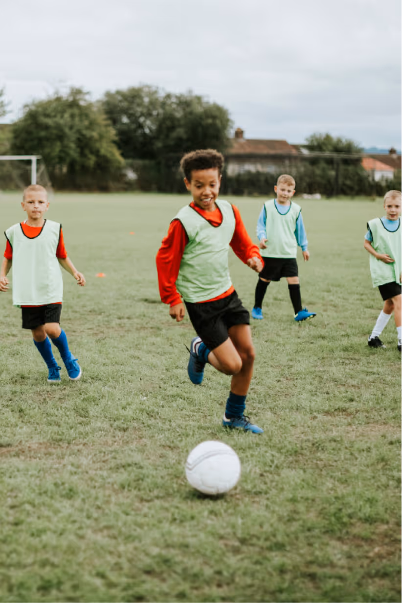 Young boys playing soccer on a grassy field, one boy in a red shirt and green vest running towards a white soccer ball.