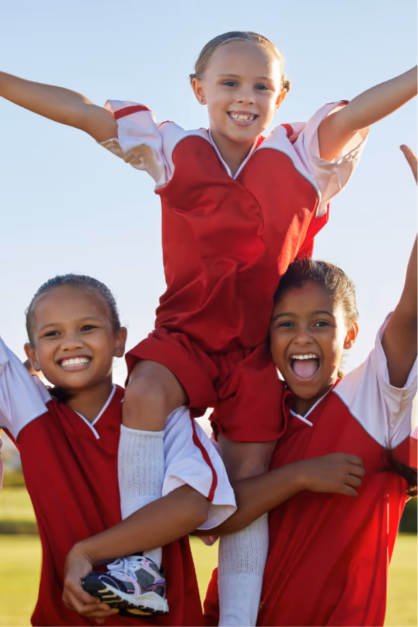 Three smiling children in red and white soccer uniforms celebrating outdoors, with one child lifted on the shoulders of the other two.
