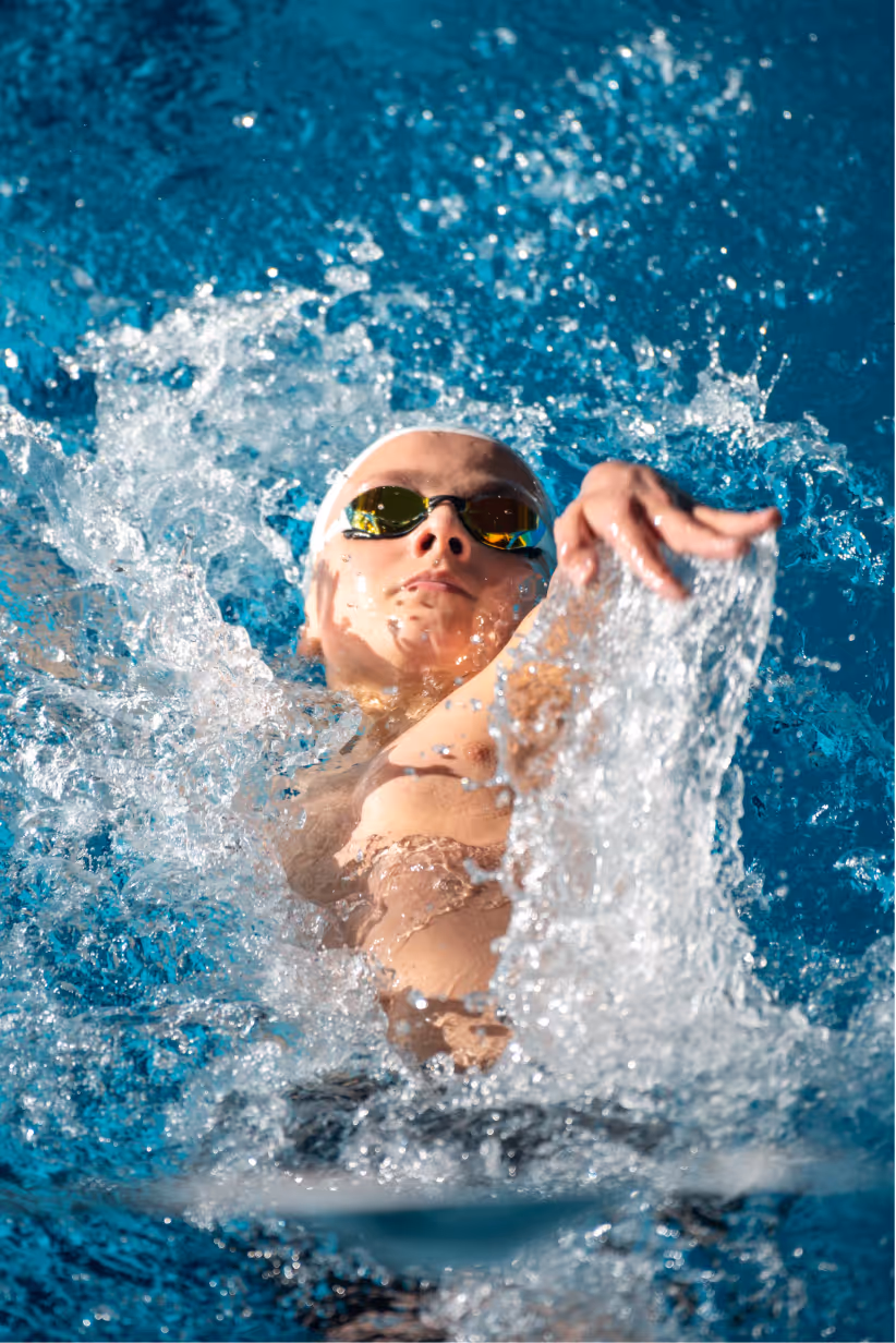 Swimmer wearing goggles and swim cap performing backstroke in a pool with water splashing around.