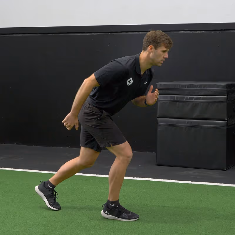 Young man in black athletic wear in a sprint start position on green turf indoors.