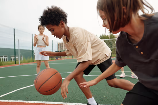 Two boys playing basketball and dribbling on an outdoor court with a girl watching in the background.