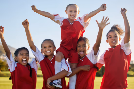 Five joyful young girls in red and white soccer uniforms celebrating on a grassy field with one girl raised on others' shoulders.