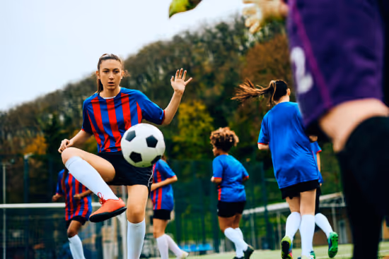 Young female soccer player in red and blue striped jersey controlling a soccer ball on a field with other players in the background.