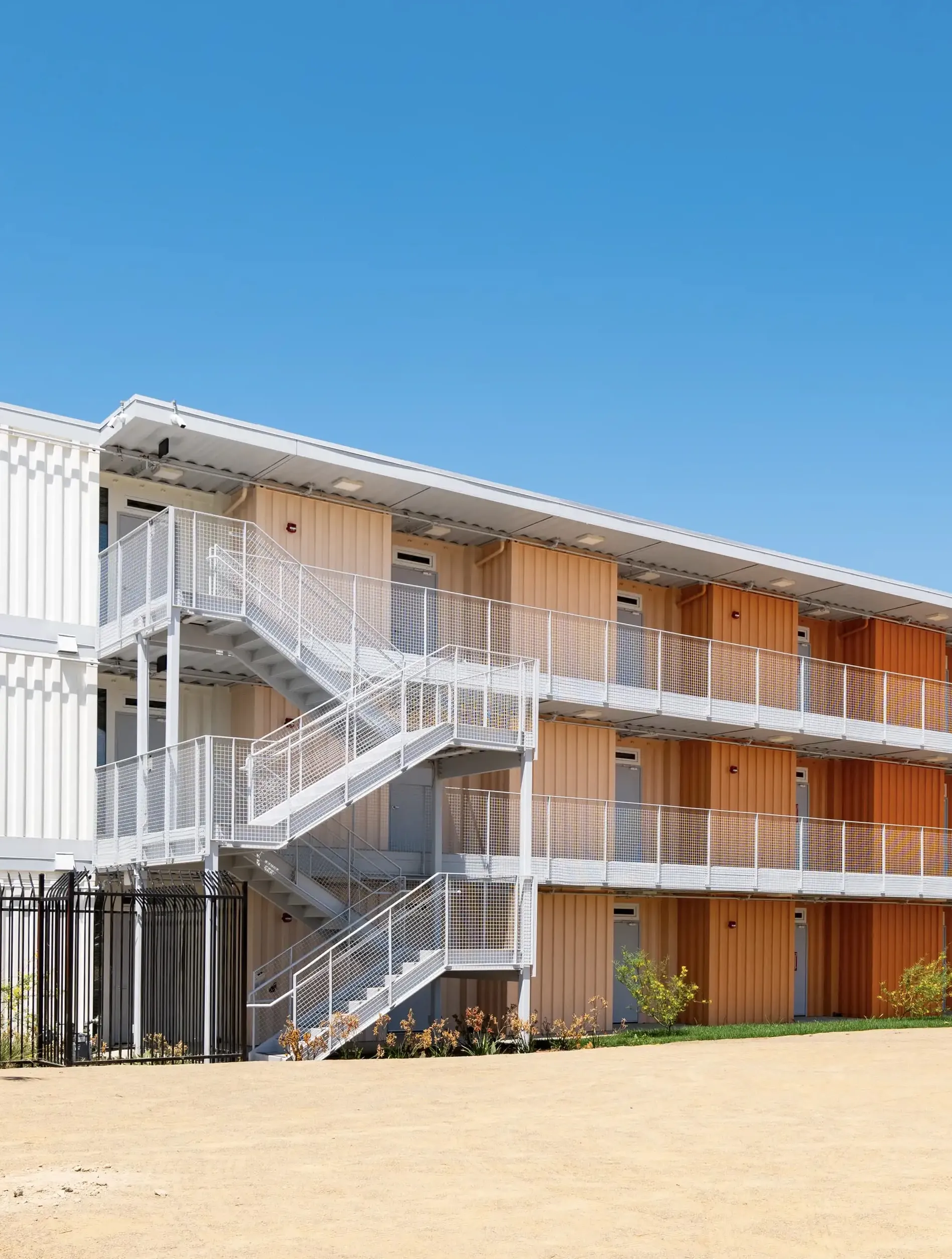 Three-story apartment building with exterior white metal staircases and peach-colored panels under a clear blue sky.