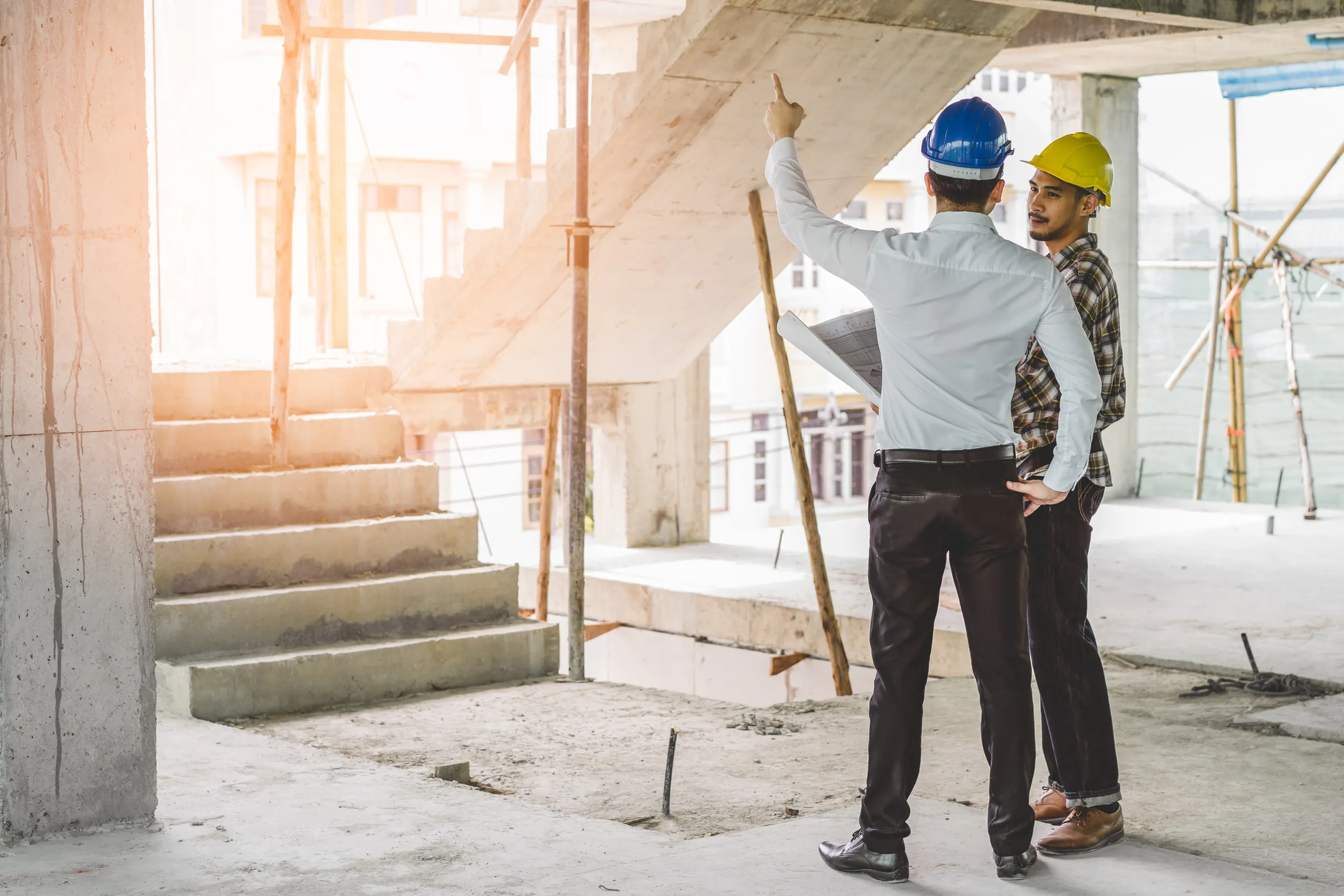 Two construction workers wearing hard hats discussing plans inside an unfinished building with concrete stairs.