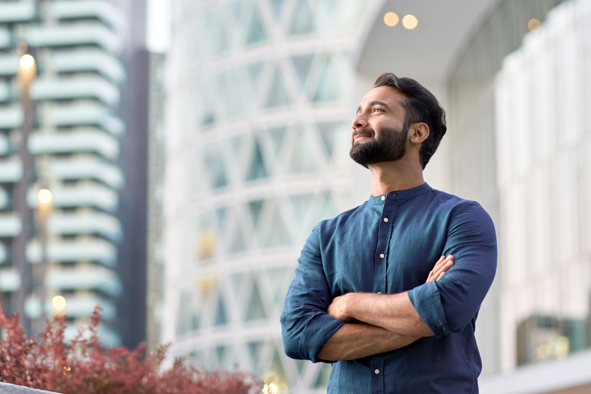 Confident man with folded arms standing outdoors with modern buildings in the background.