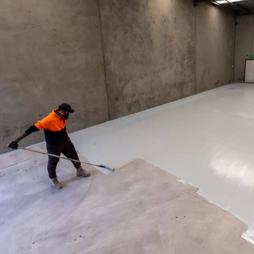 Worker in an orange hoodie applying epoxy coating to a large concrete warehouse floor with a roller.