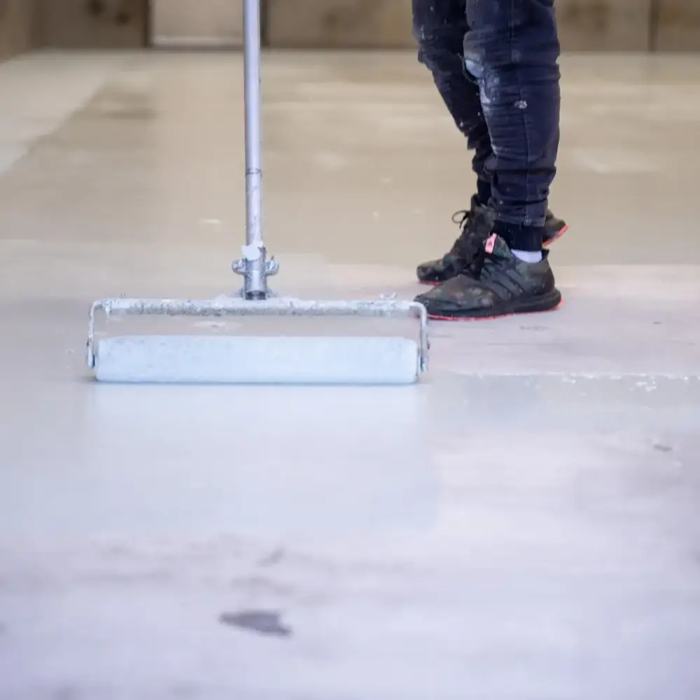 Close-up of a worker applying epoxy coating on a concrete floor using a roller, with freshly coated and uncoated sections visible.