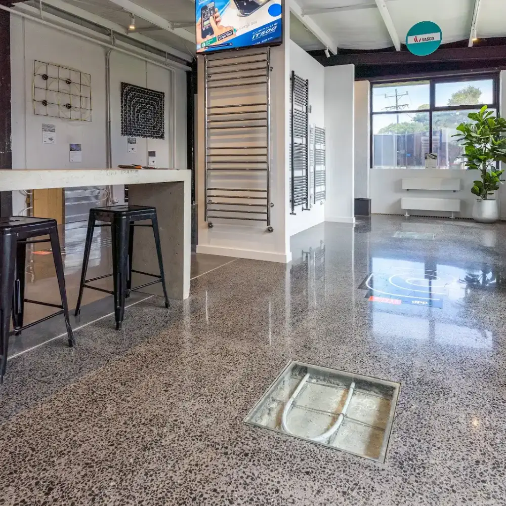 Showroom interior with polished terrazzo flooring, bar stools, wall-mounted heating panels, and a glass floor panel revealing pipes.
